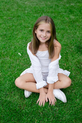 Fototapeta premium pretty tween girl in white clothes sitting on green grass outside in park on sunny summer day. charming ukrainian child portrait