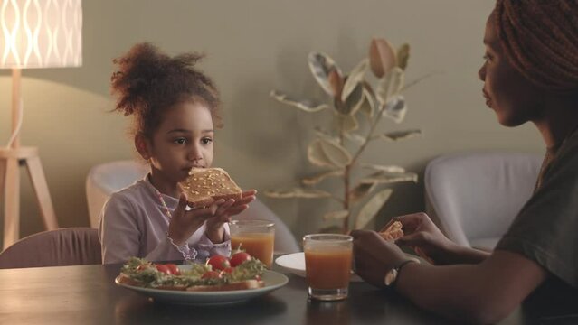 Waist up slowmo of young Black woman with dreadlocks and her 6 year old daughter eating homemade sandwiches at kitchen table and having conversation, spending leisure time together