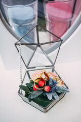 Decorative glass gift box for a wedding, on a light background. Inside are gold wedding rings for the bride and groom, as well as orange-pink flowers and green leaves.