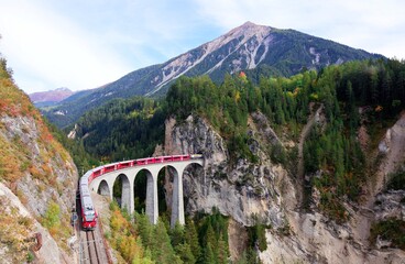 A local train coming out of the tunnel in a vertical cliff  crossing the famous Landwasser Viaduct over a deep gorge with fall colors on the rocky mountainside in Filisur, Grisons, Switzerland