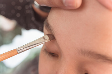 Closeup of a MUA or Makeup artist applying brow filler with a slanted eyeliner brush on the eyebrow of a young female asian model. Early stages of makeup. - obrazy, fototapety, plakaty