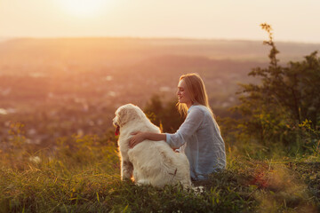 A young woman and her dog sitting on the hill in the countryside on a summer evening. A lady and her faithful friend enjoying beautiful landscape at summer sunset.