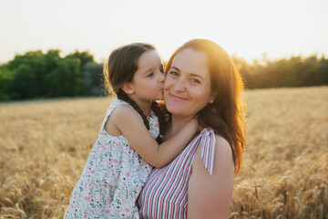 Little child daughter hugging and kissing mother while standing in the middle of the wheat field.