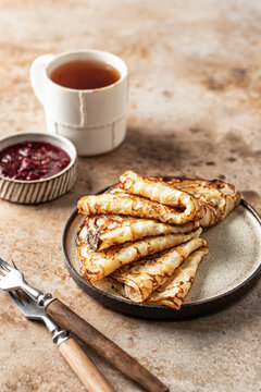 Crepes in brown plate. Breakfast composition with blinis, forks, tea, jam on brown background