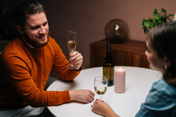 High-angle view of smiling bearded young man talking celebration toast and clinking white wine glass with girlfriend during romantic dinner. Love couple celebrating anniversary or Valentines day.