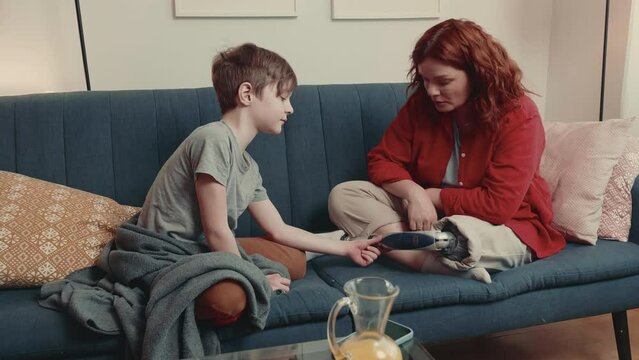 Happy curious Caucasian teen age boy touching mothers bionic prosthetic leg talking, sitting together on couch at home.