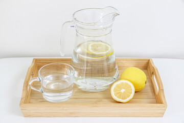 Refreshing and healthy natural citrus lemon flavoured water, lemonade, in glass jug and cup, besides full and sliced lemon, in a wooden tray, on clean white table