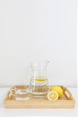 Refreshing natural lemon water, lemonade, in glass jug and cup, besides full and sliced lemon, in a wooden tray, on white table, against white wall