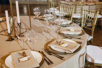 On the table in wedding banquet area are gold plates, napkins, cutlery, glasses, there are candles, menu cards lie on the plates.
