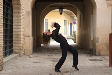 Young man with beard and long hair, wearing black transparent shirt with black polka dots and red roses, black pants and jacket, dancing flamenco in the city. Concept art, dance, culture, tradition.