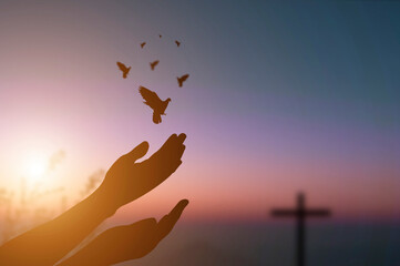Silhouette hand of woman praying and free bird enjoying nature on sunrise and orange background with sunlight with a cross in the background.