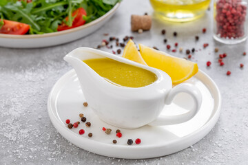 Salad dressing with oil, lemon juice, salt and pepper served in white ceramic saucepan. Horizontal, ingredients and plate with arugula and tomato salad on background. Vinaigrette or Citronette sauce.