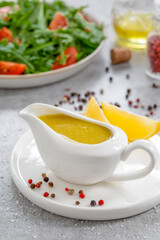 Salad dressing with oil, lemon juice, salt and pepper served in white ceramic saucepan with ingredients and plate with arugula and tomato salad on background. Vinaigrette or Citronette sauce. Vertical