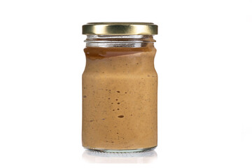 Close-up of mustard in a transparent glass jar with a closed gilded metal lid on a white background.