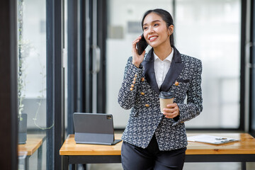 Smiling Happy Asian businessman standing and talking on a smartphone holding a coffee cup in an office
