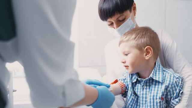little boy is taking blood from a vein for analysis. The child sits in the arms of his mother in the manipulation room of the clinic.