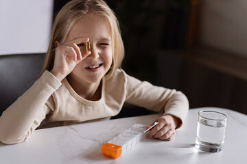 Little Caucasian Girl in pajamas taking natural vitamin omega-3 from plastic box on kitchen at home early morning. Adorable child Takes supplements. - obrazy, fototapety, plakaty