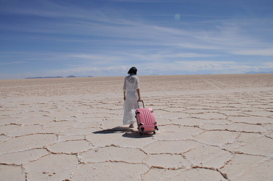 Rear view of lonely woman in white dress and with pink suitcase walking into the distance of Salar de Uyuni salt flat, Bolivia
