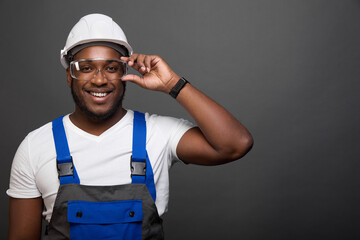 Portrait of African male engineer architect dressed in special uniform and protective helmet, adjusts his glasses with his hand. Worker stands smiling against gray wall in overalls with blue straps - obrazy, fototapety, plakaty