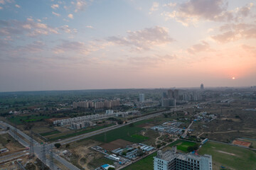 Naklejka na ściany i meble aerial drone shot passing over a building with homes, offices, shopping centers moving towards skyscapers in front of sunset showing the empty outskirts of the city of gurgaon