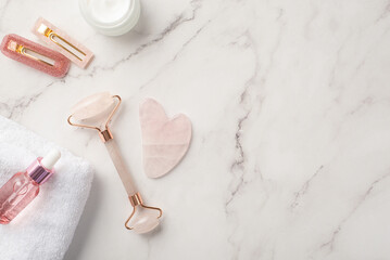 Skincare concept. Top view photo of rose quartz roller gua sha massager cream jar pink glass transparent bottle stylish barrettes and white towel on white marble background with copyspace