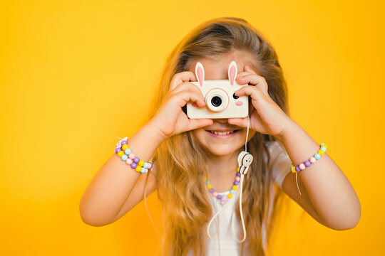 Portrait of a cute little blonde girl takes a picture with a child's camera, poses for a photographer on a yellow background