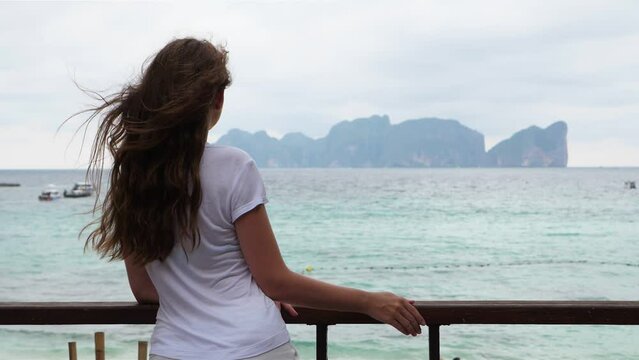 Woman turn head and look to tropical island, long hair fly in air, view from behind. Cloudy and windy weather at Phi Phi Don. Dreamy tourist want to reach famous Koh Lee islands