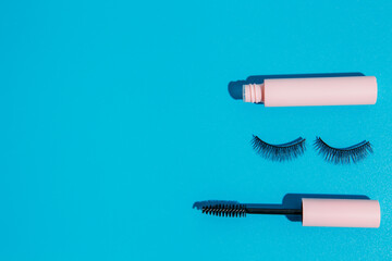pink clear mascara brush lies next to an open tube and false lashes on a blue background, copy space. Makeup cosmetics set. Flat lay, top view.