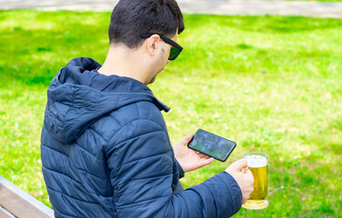 young man is watching football at the mobile phone and holding a glass of beer in another hand, in...