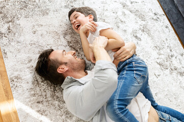 Happy father and school-age son spending time together at home, have fun lying on the floor in cozy...
