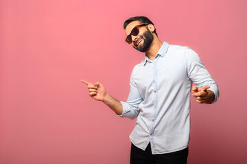 Overjoyed Indian man wearing sunglasses dancing, makes movements to music, smiles positively, being...