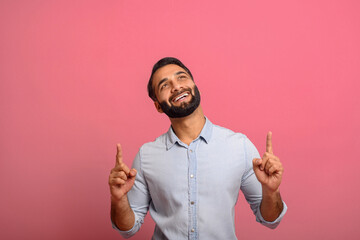Cheerful overjoyed Indian guy points fingers up at empty copy space, isolated on pink background....