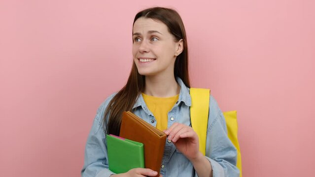 Portrait of confused fun girl teen student hold books spreading hands say oops ouch oh omg i am so sorry, wears shirt and yellow backpack, posing isolated over pink color background wall in studio