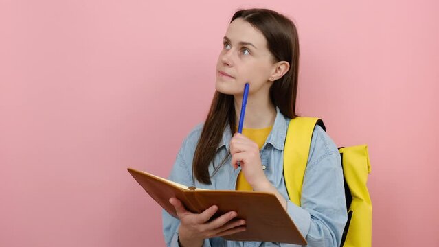 Portrait of pensive girl teen student wears shirt and yellow backpack writing down in exercise book notes, isolated on pastel pink color background wall in studio. Education in university college