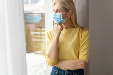 Senior mature lady in protective face mask looking at the window and feeling sad depressed and lonely during coronavirus pandemic, lockdown struggles concept