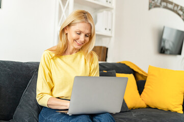 Modern mature 60s female enjoy leisure weekend at home, browsing wireless internet on computer gadget. Happy elderly woman sit relax on couch in living room, work on laptop, smiling