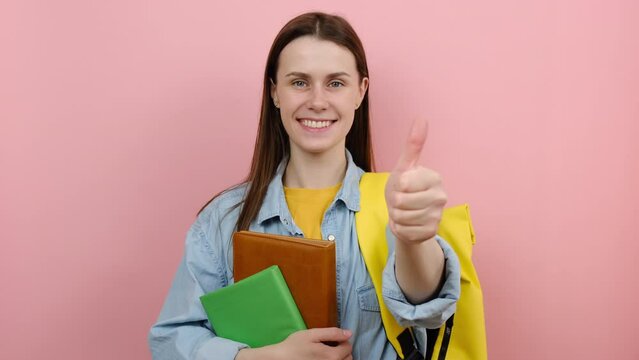 Portrait Of Happy Girl Teen Student Wears Shirt And Yellow Backpack Hold Books Show Thumb Up Like Gesture, Isolated On Pastel Pink Color Background In Studio. Education In University College Concept