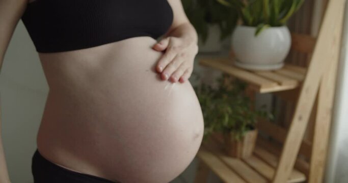 Young pregnant woman applies stretch marks massage lotion on her belly. Woman uses moisturizing cream to prevent stretch marks. Maternity prenatal care and woman pregnancy concept.