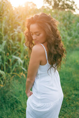 Portrait of a beautiful dark-haired curly young woman 30 years old in a white sundress in a summer field at sunset. Beautiful summer model.