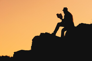 silhouette of a man in a hat reading a book sitting on a mountainside