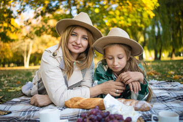 Blonde woman in hat have picnic with daughter in good autumn day. Outdoor portrait of pretty little girl spending time with mother in park.
