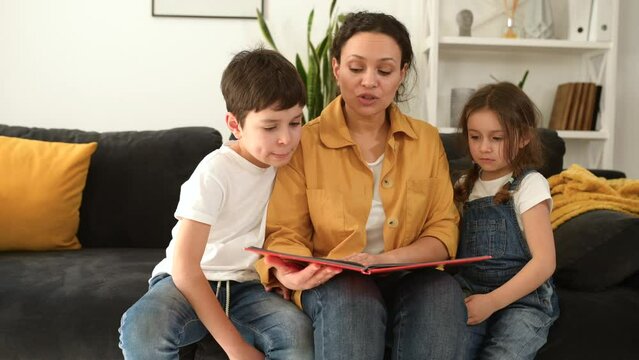 Portrait of mother and her little cute kids sitting on the sofa together, lovely mom and adorable toddler son and daughter reading book. Love and care concept