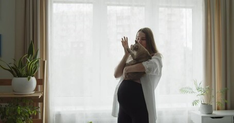 Happy cheerful pregnant woman dances with a cat in her arms near the window at home. Expectant loving mother relax at home listening to the music. Maternity prenatal care and woman pregnancy concept.