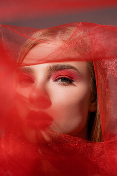 Model with red eye shadow looking at camera near blurred cloth on grey background.