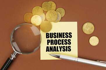 On a brown surface are coins, a pen, a magnifying glass and stickers with the inscription - BUSINESS PROCESS ANALYSIS