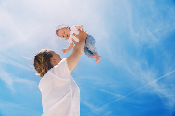 Mother throwing baby up against the blue sky. Happy family outdoors. Mom and baby at summer on...