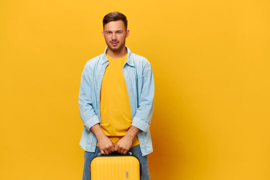 Happy Enjoyed Tanned Handsome Man In Blue Shirt Smiling At Camera Hold Suitcase Both Hands Posing Isolated On Orange Yellow Studio Background. Copy Space Banner Mockup. Trip Journeys Concept