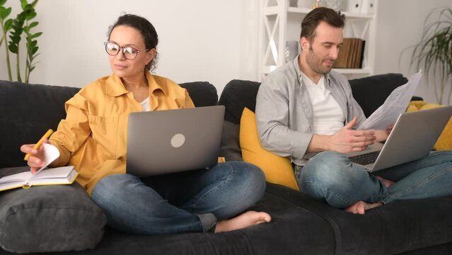 Couple using laptops for remote work on the sofa at home, focused spouses working on the distance, answering emails, doing paperworks in home atmosphere
