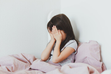 brunette toddler girl sitting in bed, covering her face with her hands, crying, frustrated