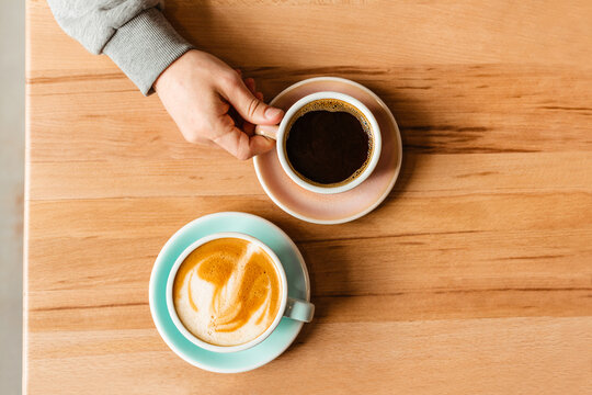 A Man's Hand Holds A Hot Americano Next To A Cappuccino. Coffee In Ceramic Cups For Two View From Above. Stylish Concept For Your Diamond And Roasted Coffee Advertising